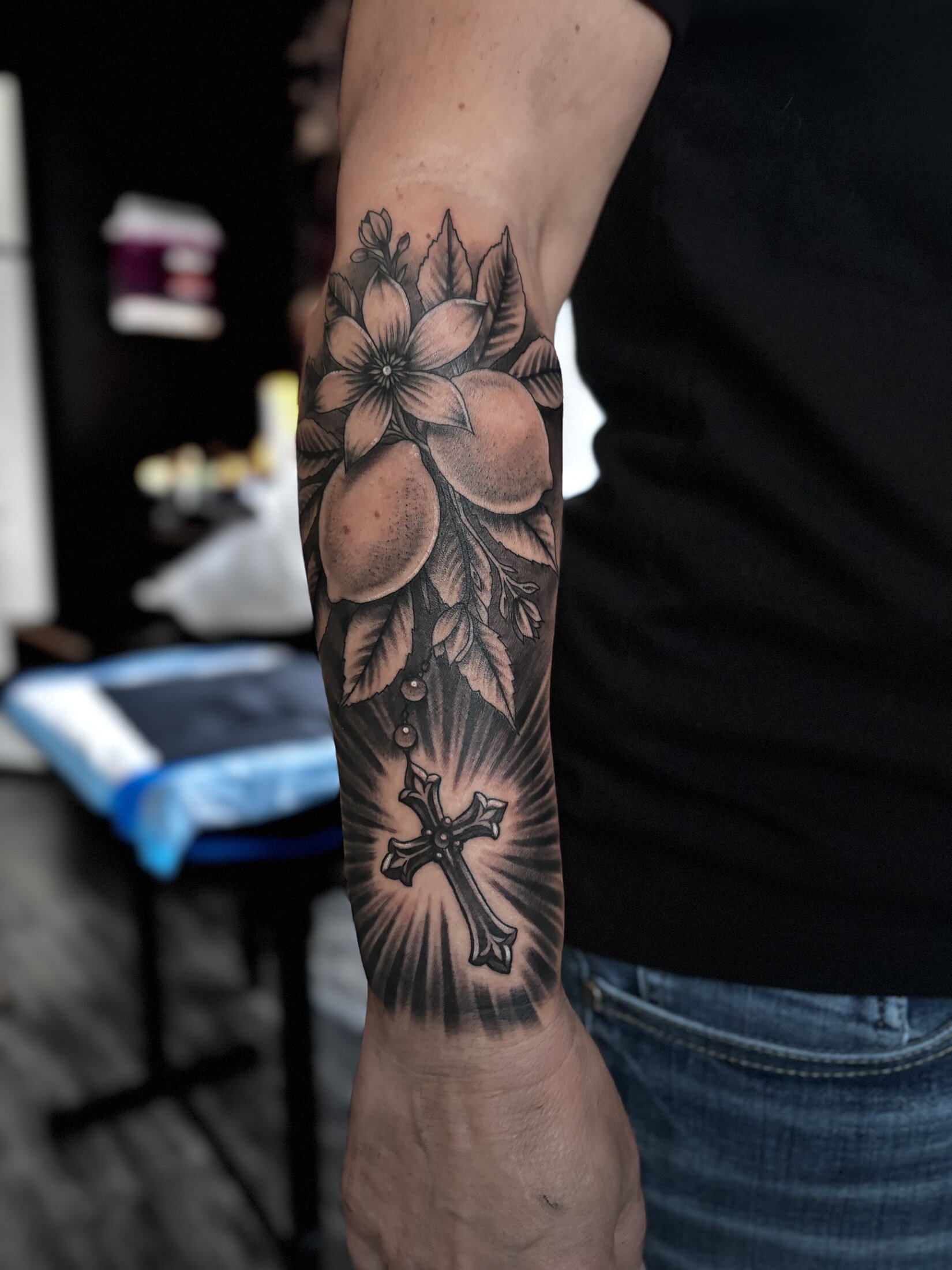 Black and grey realism tattoo of orange blossoms and a radiant cross on a forearm