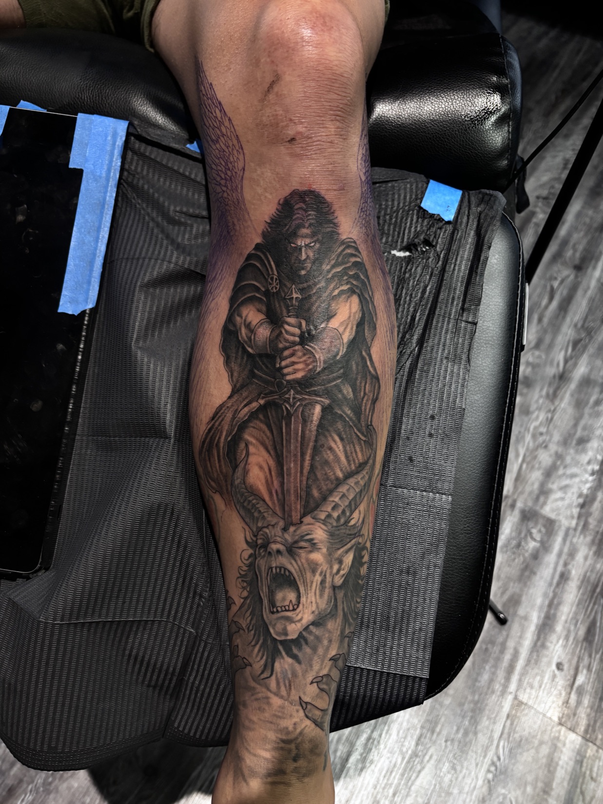 Black and grey realism tattoo of a winged guardian angel holding a sword over a screaming demon on a calf